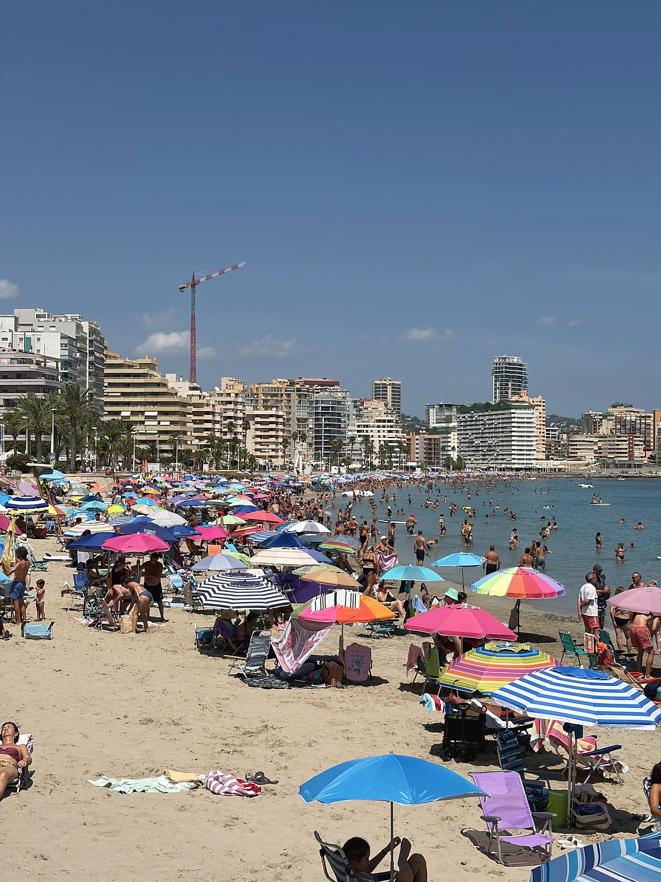 Preplnená pláž v Calpe počas hlavnej sezóny, v pozadí vežiaky a stavebný žeriav